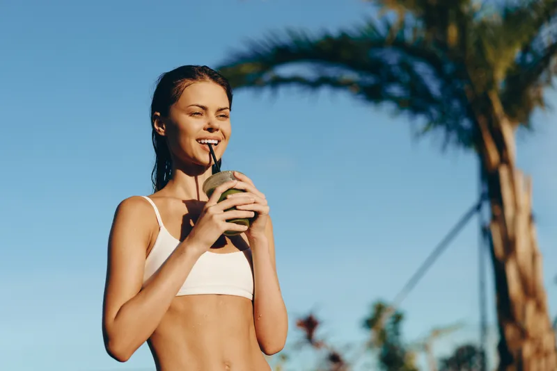 Healthy woman wearing a white sports top drinks a smoothie outdoors, smiling, sunlit scene with palm trees, capturing a carefree lifestyle and fitness inspired moment