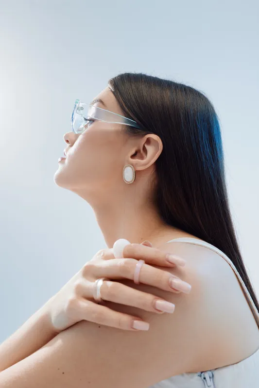 Profile of a young woman with long dark hair wearing transparent glasses and elegant earrings. She poses with crossed arms against a light background. Concept of beauty fashion.