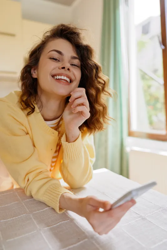 A cheerful young woman in a yellow cardigan sits at a bright home desk, smiling at her phone, capturing a relaxed, confident moment of everyday life.
