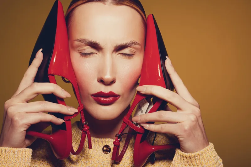 Close-up of a woman with closed eyes holding red high heels near her face. Fashion concept with bold makeup and stylish accessories on a warm brown background.