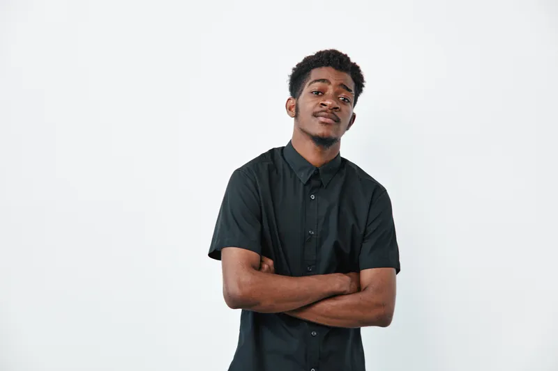 Young African American man with dark skin and short curly hair wearing black shirt standing with crossed arms against white background. Confident expression and casual pose.
