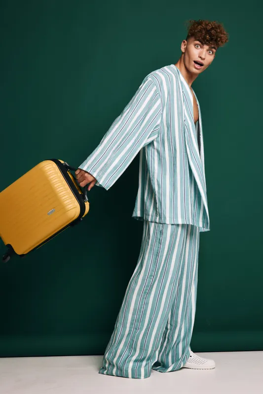 Young man in striped suit holding yellow suitcase with wheels, standing against green background, looking surprised and ready for travel or business trip.
