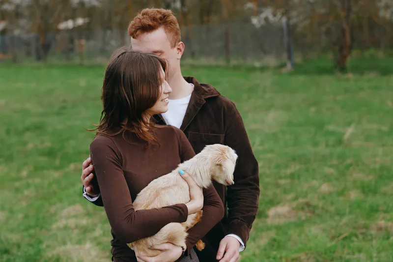 Loving couple outdoors, embracing with a gentle goat kid nestled between them, soft sunlight, casual clothing, lush field backdrop, candid connection, warm natural tones, lifestyle moment