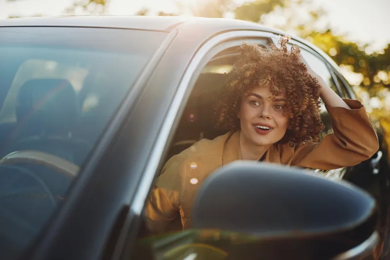 Smiling woman with curly hair sits in a car with the window down, sunlight bathes a retro lifestyle portrait, candid and warm, capturing a joyful road moment colorful relaxed atmosphere emphasizes