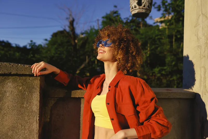 Curly haired woman wearing a red jacket and yellow crop top leans on a stone railing outdoors in bright sun, wearing sunglasses and a relaxed confident smile, a bold fashion moment