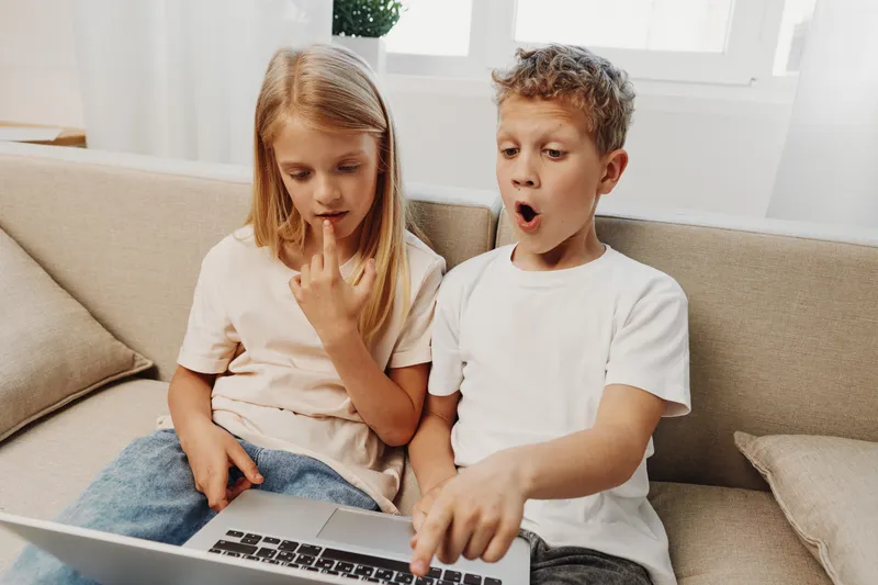 Curious children sitting on a sofa looking surprised at a laptop screen. Boy points excitedly while girl watches thoughtfully in a bright cozy room. Concept of discovery and learning.