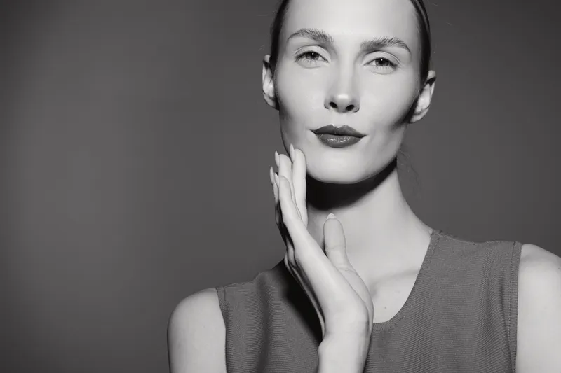 Portrait of a young white woman with pale skin and red lips in black and white, touching her face with hand in studio light.