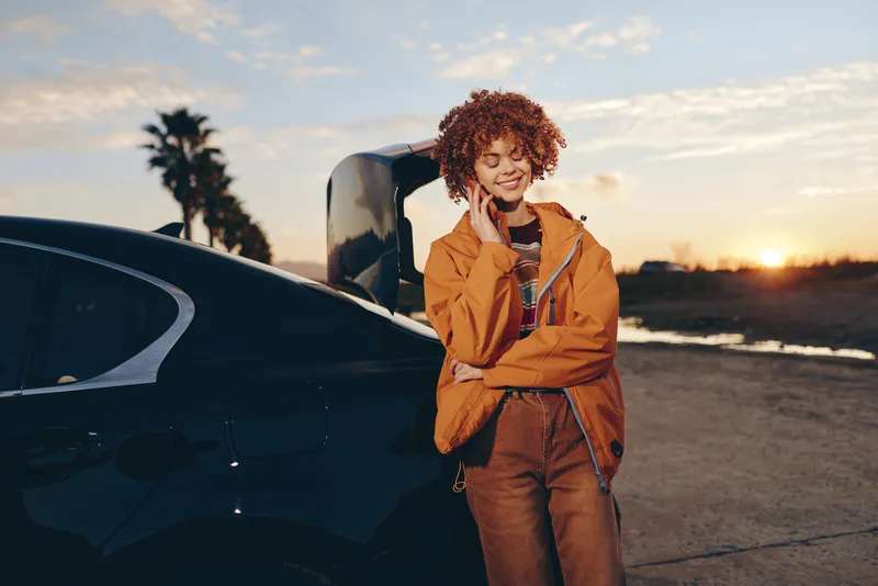 Woman lifestyle outdoor portrait at sunset near car at beach lot, curly hair, orange jacket, relaxed smile, leaning beside vehicle, travel vibe in warm golden light with evening sky