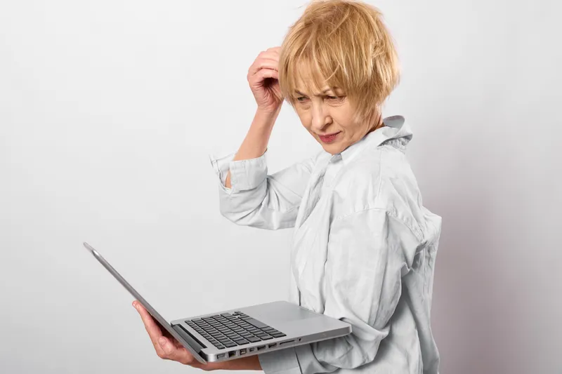 Stressed elderly woman in white shirt holds a laptop, rubbing temple with headache and worried face, plain studio background, concept of stress, burnout, anxiety, remote work workplace