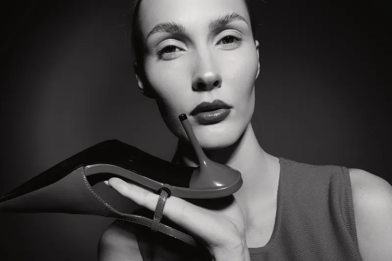 Close-up of a fair skinned Caucasian woman holding a high heel shoe, wearing sleeveless top. Fashion concept in monochrome studio.