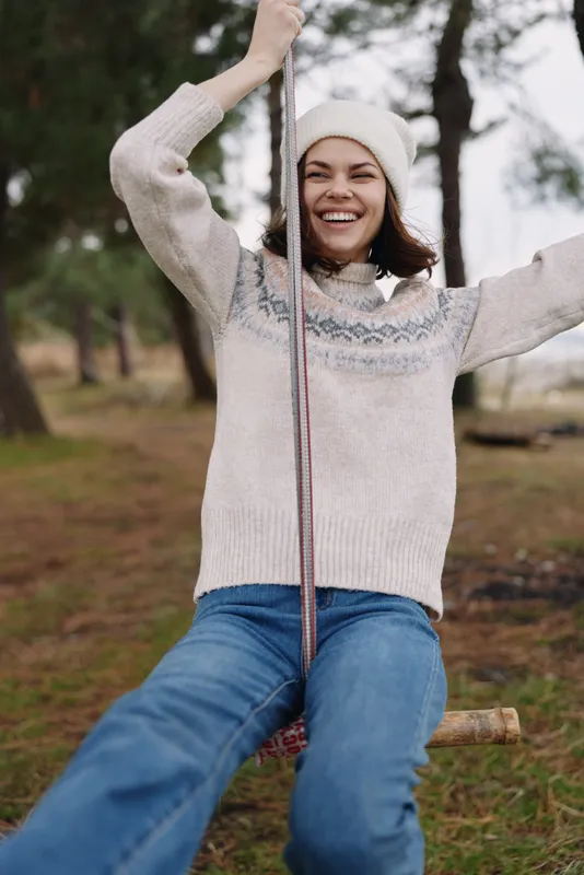 Girl on a rope swing in a park, smiling, wearing a chunky sweater and beanie, enjoying outdoor play. Sunny day with trees in the background, casual and joyful.