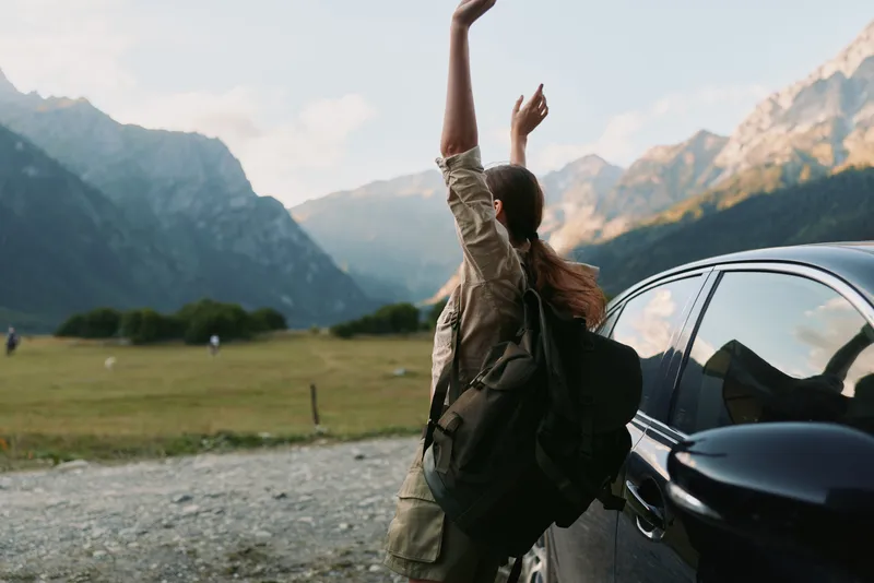 Young woman with backpack stands beside a car in a mountain valley, arms raised in a joyful, liberated moment after a scenic hike under clear blue skies.
