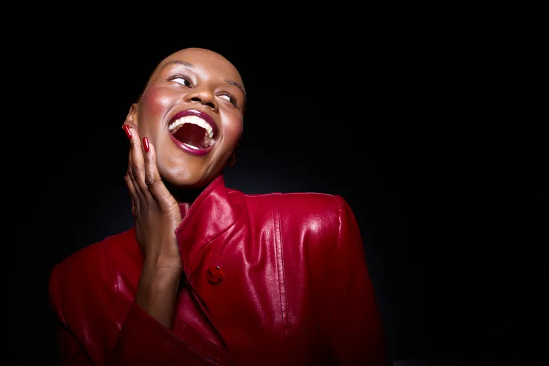 Joyful black woman with dark skin wearing a red leather jacket laughs with hand on face against a black background. Concept of happiness, fashion, and confident female beauty.