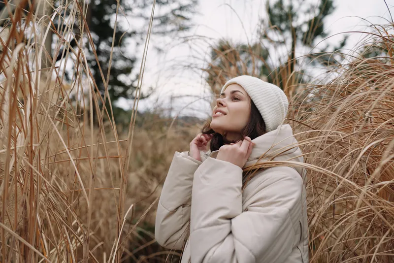 Woman wearing a beige coat and white beanie stands among tall dried grasses in a winter landscape, calm and thoughtful pose, natural light highlighting texture and serenity in an outdoor setting