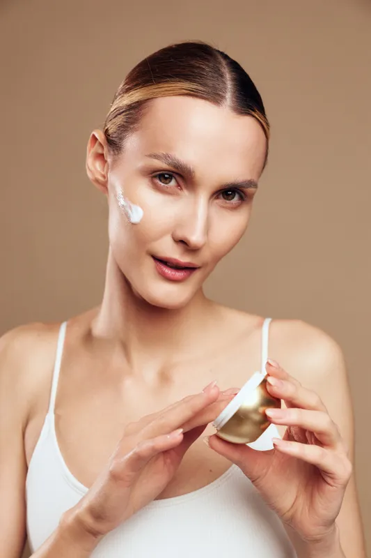 Young woman applies face cream on cheek holds jar with moisturizer looks at camera wears white tank top isolated on beige background skincare beauty concept.