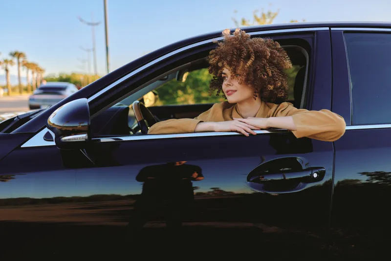 Casual woman leaning on a car window with curly hair. Outdoor daytime scene with warm retro film color tones. Lifestyle portrait conveying ease, freedom, and self expression.