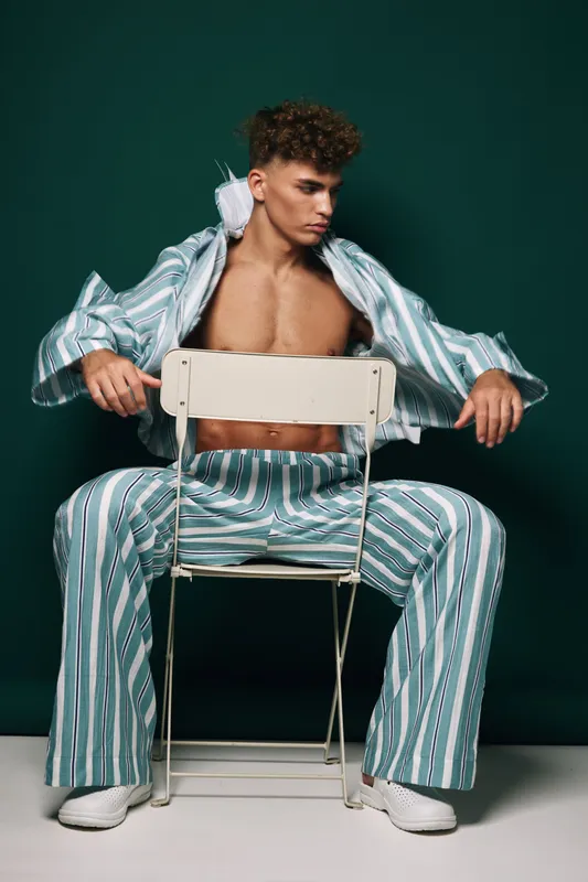 Young man with curly hair wearing striped pajamas sitting on chair backwards against dark background. Casual fashion, relaxed pose and modern style with white sneakers and open shirt.