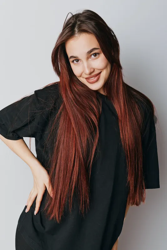 Young woman with long red hair smiling and posing in casual black t shirt on light background. Portrait of happy female with natural makeup and confident expression indoors.