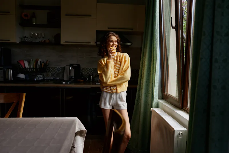 Woman stands by a sunlit window in a cozy home kitchen, wearing a yellow sweater and white shorts, thoughtful pose, natural light, relaxed candid moment, modern casual living space