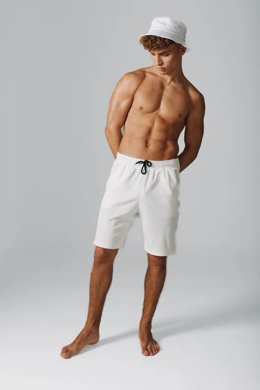 Young shirtless man posing with white bucket hat and white shorts on grey background. Casual summer fashion with fit muscular body and shorts posing barefoot. Trendy youth and style studio shoot.