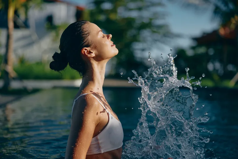 Relaxed woman in a white bikini enjoys a pool splash, sunlit water droplets, and a calm outdoor moment, capturing summer leisure, wellness, and self care with a bright joyful mood.