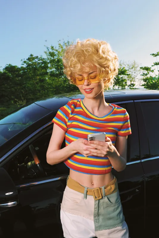 A retro lifestyle portrait of a woman with curly blond hair in a striped crop top, standing beside a car and looking at a phone in bright outdoor light.