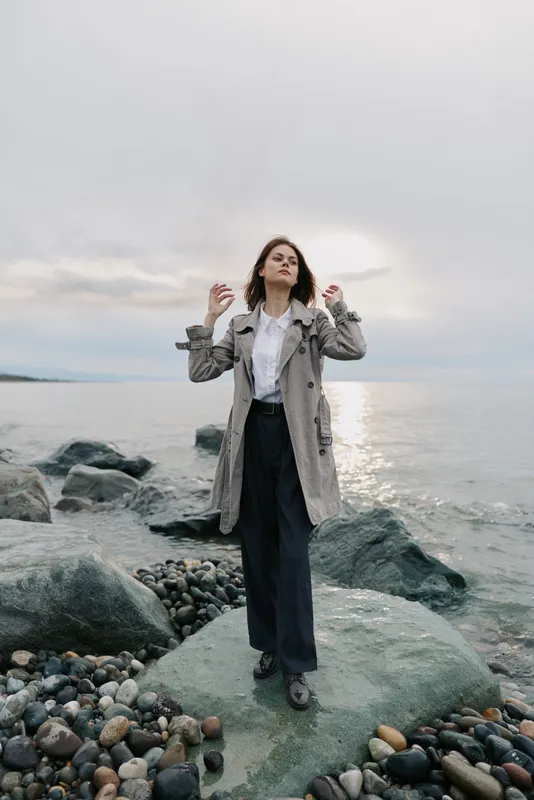 Woman in long coat stands on rocky shore near calm sea under cloudy sky. She looks confident and relaxed, capturing a peaceful moment by the water at sunset.