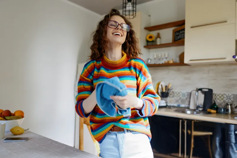 A cheerful woman in a multicolored striped sweater holds a blue cloth in a bright kitchen, smiling broadly. A candid scene of home cleaning and relaxed lifestyle.