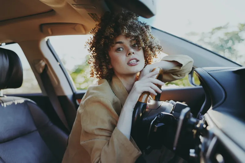 A stylish woman sits behind the steering wheel in a car, casual attire, driving, looking toward the camera with curiosity. The bright interior and sunlit scene convey a modern, relaxed lifestyle.
