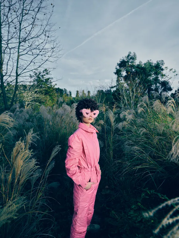 Pink jumpsuit clad woman stands in tall grass outdoors with heart sunglasses, creating a playful fashion portrait amid a calm field and blue sky, showcasing bold color and relaxed confidence.