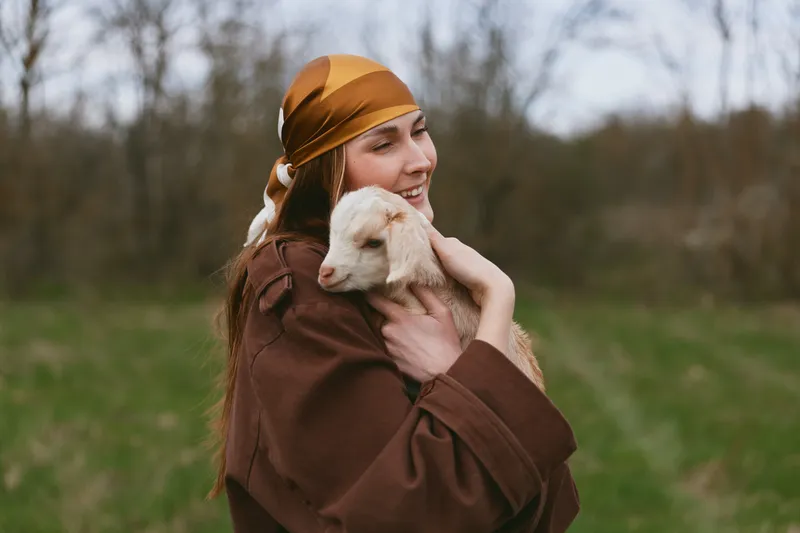 Warmhearted woman cuddling a goat kid outdoors in a field, capturing a tender bond with nature. Rustic autumn setting, soft light, lifestyle moment of care and joy.