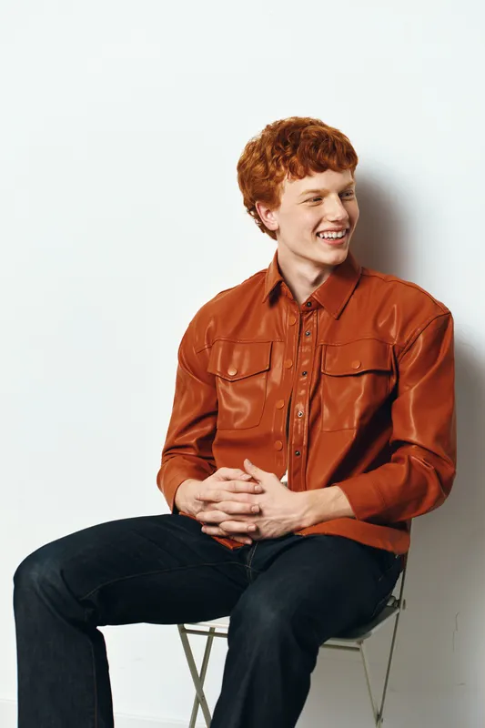 Young man with red hair sitting on chair smiling and looking sideways. Casual style with brown shirt and dark jeans on white background in studio.