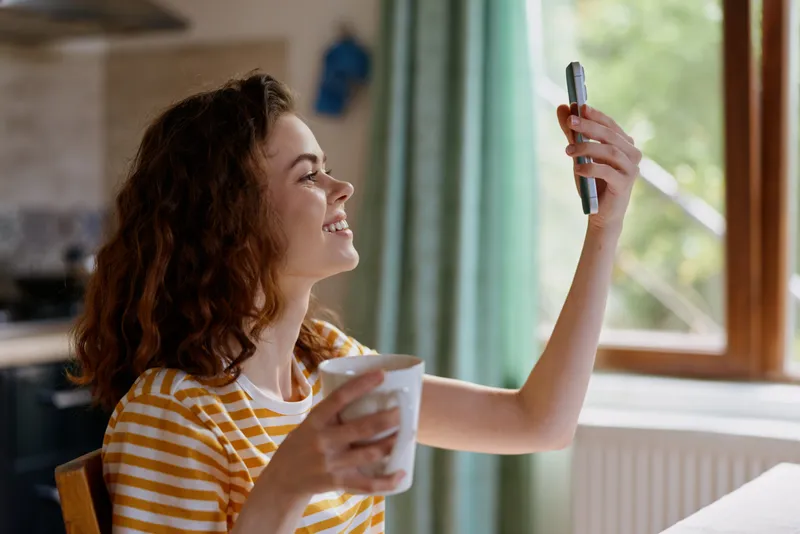 Joyful woman at home indoors taking a selfie by the window, holding a coffee mug, sunlit moment, striped shirt, relaxed smile, natural colors, cozy living space