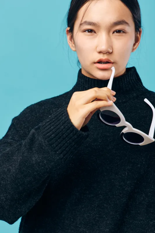 Young woman in black sweater holding white sunglasses with one hand near face on blue background. Portrait, fashion, casual style and confident expression for modern lifestyle.