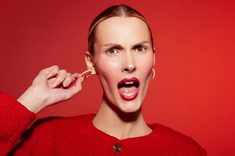 Angry woman in red sweater and earrings pulls her ear with open mouth on red background. Concept of frustration, irritation, and emotional expression in studio portrait.