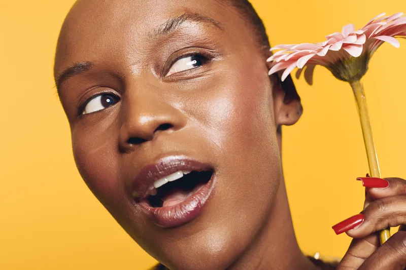 Close-up of a happy dark skinned African woman holding a pink flower near her ear on a bright yellow background. Concept of beauty, joy, nature, and natural skincare.