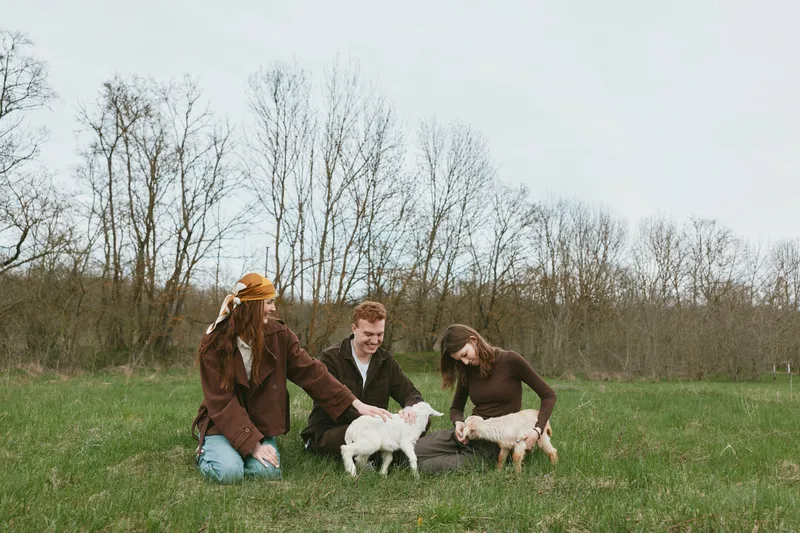 Three young adults enjoy a casual moment together in a grassy field playing with two baby goats during an overcast day in early spring outdoors.
