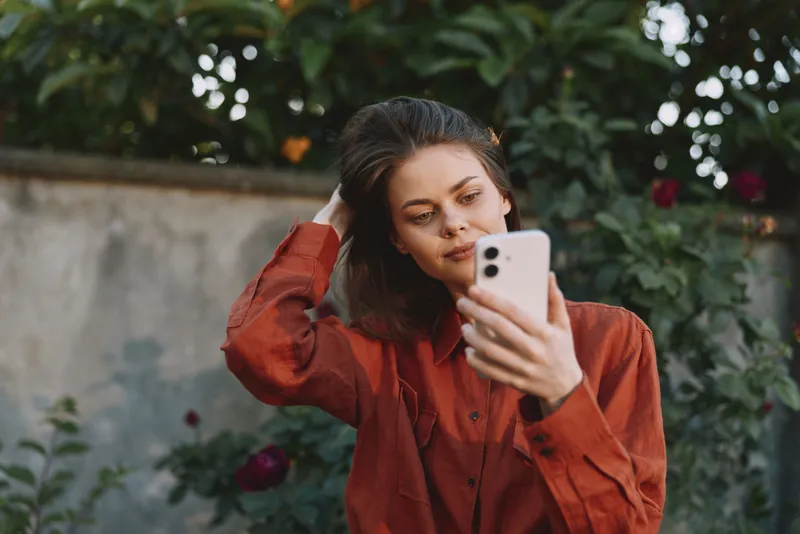 Woman lifestyle portrait using smartphone outdoors in a garden, wearing a red shirt, thoughtful pose, natural light, calm mood, leafy background, candid everyday moment