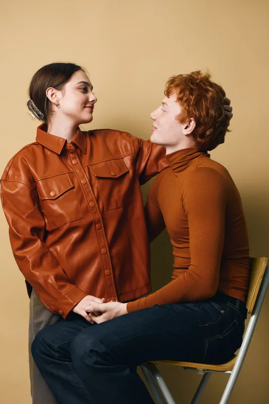 Couple, love, affection and togetherness with woman in leather jacket and man in brown turtleneck sitting and looking at each other against beige background.