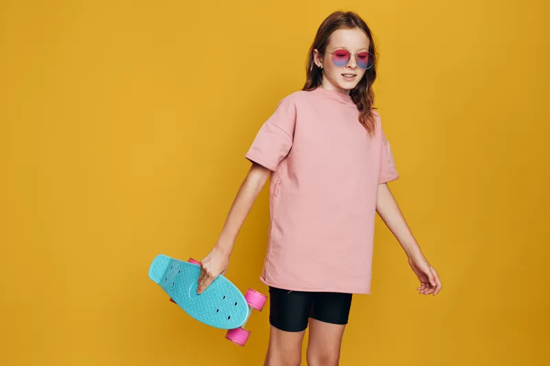 Young girl in pink shirt and sunglasses holding blue skateboard standing on yellow background. Casual style and confident pose.