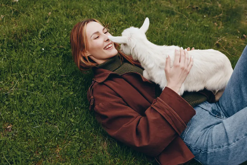 Relaxed young woman lying on grass, softly cuddling a white goat kid, enjoying a gentle outdoor moment, casual jacket and jeans, warm sunlight, peaceful countryside lifestyle scene.
