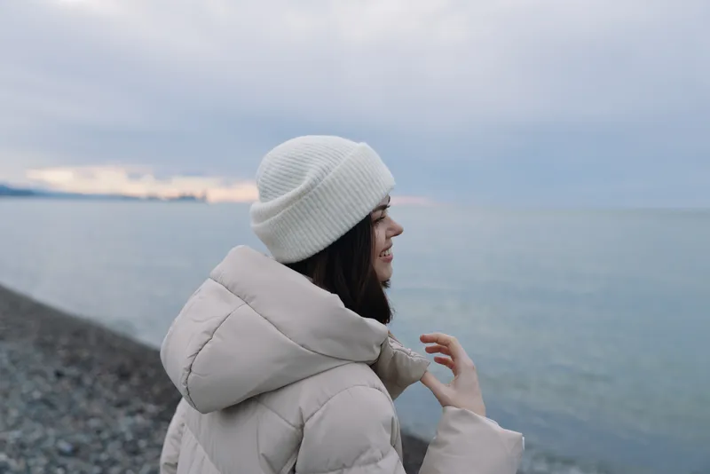 Woman by the coast in a puffy winter coat and knit beanie gazes at the sea, calm and thoughtful, with muted light and open shoreline in a simple outdoor portrait