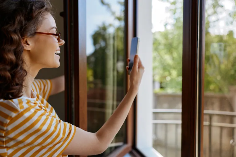 Cheerful woman indoors by a window, smiling while capturing a photo, casual striped shirt, warm sunlight, relaxed home moment, everyday lifestyle, natural lighting