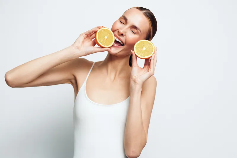 Happy woman in white tank top holding two orange halves near her face with closed eyes on white background. Concept of healthy lifestyle and fresh fruit enjoyment.