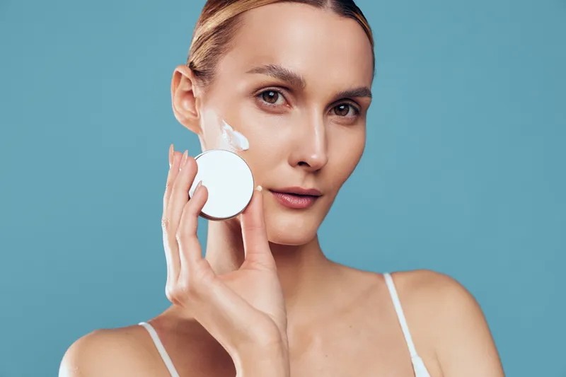 A young woman with clear skin applies white cream to her cheek using a round cosmetic sponge against a solid blue background. Concept of skincare, beauty, and facial care routine.