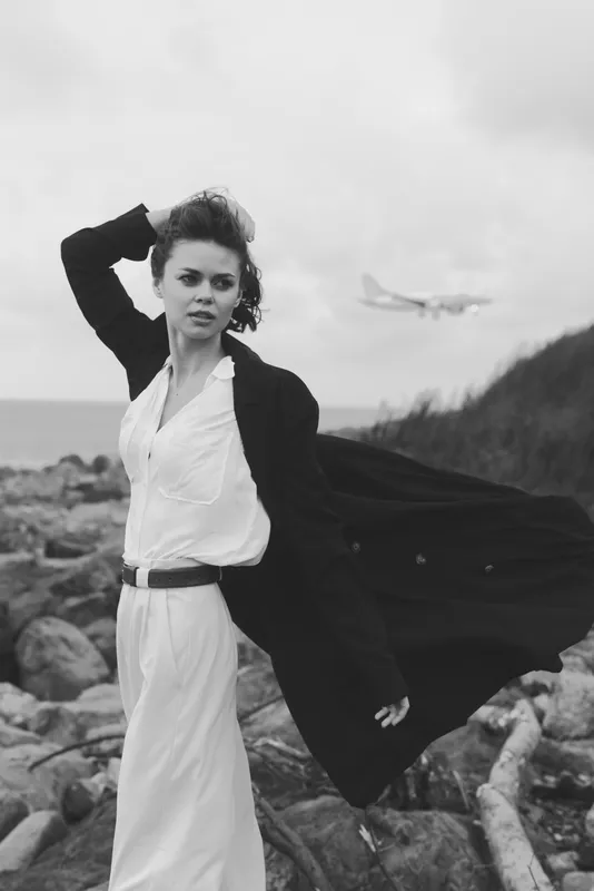 Elegant woman in a flowing coat and white dress on a windy rocky beach, distant airplane in view, showcasing fashion, movement and outdoor mood