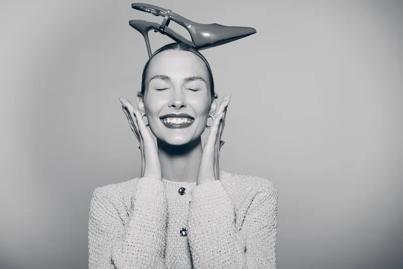 Smiling white woman with pale skin wearing a sweater and a high heel shoe on her head, eyes closed in joyful expression against a plain background.