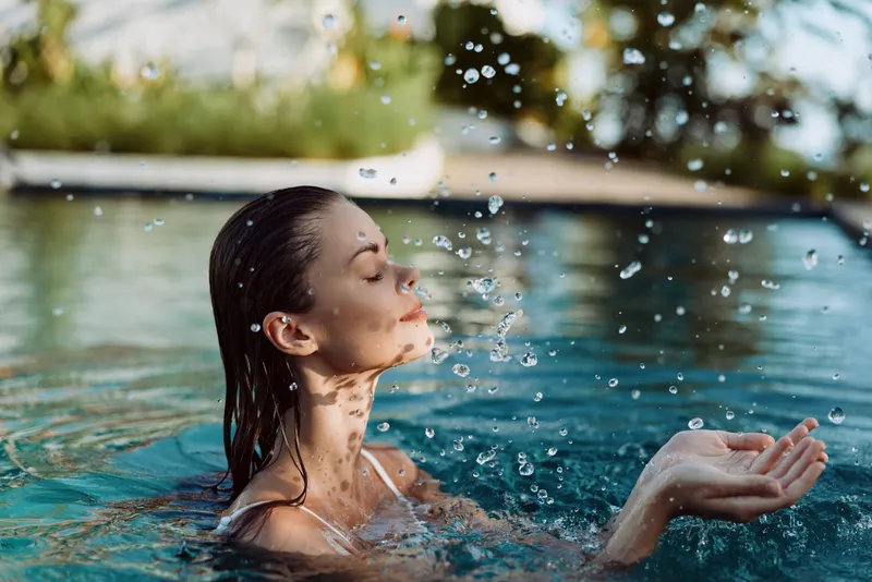 Young woman with wet hair enjoying water splash in outdoor pool. Relaxed lifestyle concept with natural light and blurred green background. Close-up on peaceful face and hands.