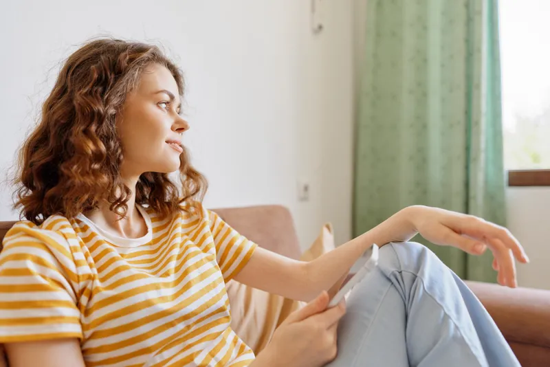 Woman with curly hair sits on a cozy couch at home, wearing a striped t shirt, enjoying a calm, relaxed moment by the window, showcasing casual lifestyle indoors.
