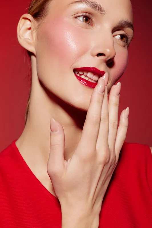 Close-up of a smiling woman with red lipstick and glossy skin touching her face. Red background and clothing create a vibrant, elegant beauty concept with natural makeup and glow.